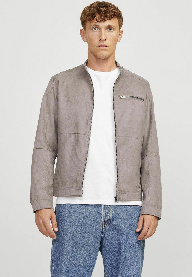 Giacca in suede grigia con tasca frontale zip, scollo rotondo e maniche lunghe, abbinata a una camicia bianca e jeans blu.
