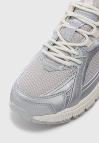 Lysegrå sneakers med mesh-overdel og sølvfargede syntetiske detaljer, flate hvite snører og en teksturert såle. Logo synlig på siden.