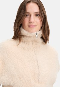 Lichtbeige fleece pullover met een hoge kraag, halve rits en een pluizige textuur. Heeft een zachte, gezellige uitstraling en minimale details.