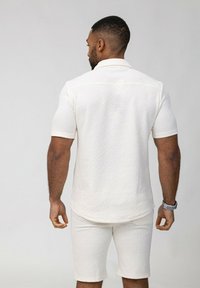Polo shirt blanc à manches courtes avec un motif géométrique texturé, associé à un short assorti. Le tissu présente un effet de motif en relief subtil.
