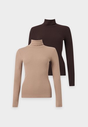 Jeune femme debout en jean bleu, col roulé beige et chaussures noires, une main dans la poche sur fond blanc.