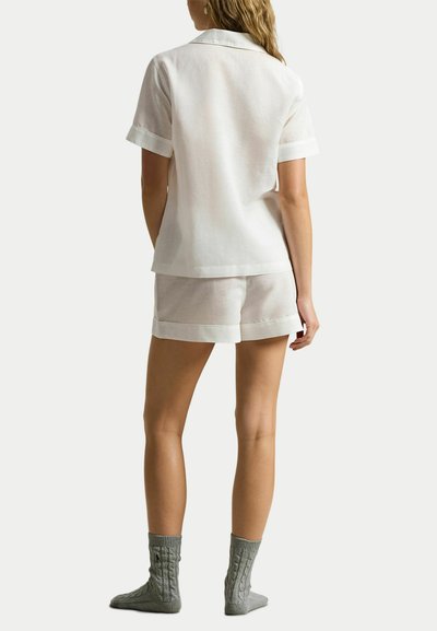 Vrouw staand met haar rug naar de camera, gekleed in een wit shirt met korte mouwen, witte shorts en grijze gebreide sokken op een effen achtergrond.