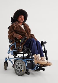 Nagy afro frizurájú személy, barna prémkabátban és fémes csizmában ül egy kék elektromos széken egy fehér háttér előtt.