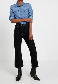 Camisa de denim azul claro con botones frontales, jeans negros de alta cintura con dobladillo deshilachado y botas de tobillo negras de punta afilada.