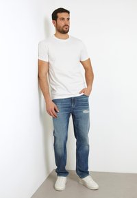 Camiseta blanca de manga corta, jeans azules desgastados y zapatillas blancas. Pose de pie contra una pared blanca lisa, con las manos en los bolsillos.