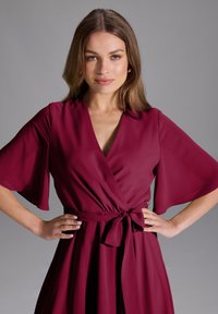 Robe portefeuille bordeaux avec des manches courtes et larges et une taille à nouer. Fabriquée en tissu lisse, elle présente un décolleté en V profond et une silhouette fluide.