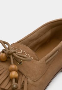 Mocassin en daim marron avec des détails en franges, un laçage décoratif et des accents en perles en bois. Caractérisé par un bout rond et un intérieur rembourré.
