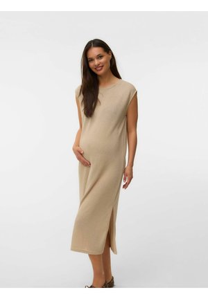 Femme enceinte aux longs cheveux foncés souriante, portant une robe midi beige sans manches avec une fente latérale, debout devant un fond uni.