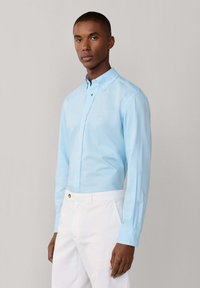 Homme portant une chemise bleu clair à boutons, manches longues, et un pantalon blanc, debout devant un fond uni gris clair.
