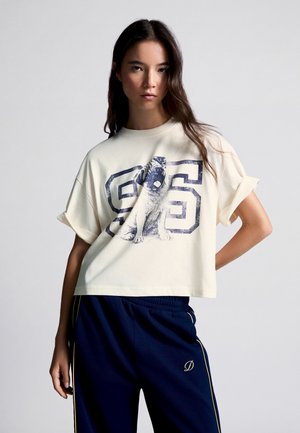 Jeune femme aux longs cheveux foncés porte un T-shirt clair et ample avec un carlin et le numéro 96, associé à un pantalon bleu foncé à bordure jaune.