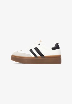 Zapatilla blanca con rayas negras, con punta redondeada y suela gruesa de goma marrón. Detalles texturizados en la punta y el talón.
