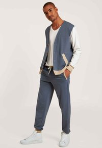 Blauwe casual jasje met beige accenten, knoopsluiting en zijzakken, gecombineerd met bijpassende blauwe joggers en witte sneakers.