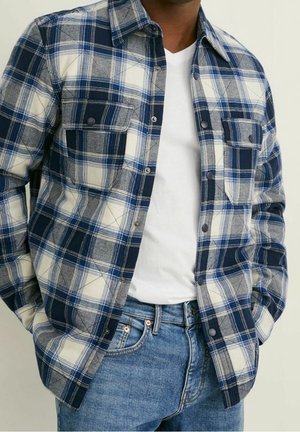 Blauw en wit geruit shirt met knoopsluiting, twee borstzakken en een kraag. Draagt boven een wit T-shirt en gecombineerd met blauwe jeans.