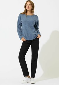 Donna in piedi con le mani in tasca, che indossa un maglione blu a maniche lunghe, pantaloni neri e sneakers bianche su uno sfondo bianco.