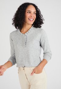 Maglione grigio a maglia con scollatura arricciata e patta con tre bottoni, abbinato a pantaloni chiari; texture morbida e maniche fitted.