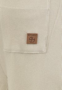 Tissu beige en maille gaufrée avec une poche carrée et une petite étiquette en cuir marron portant l'inscription "KEY LARGO" cousue sur la poche.