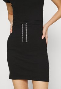 Robe noire côtelée avec un design ajusté, des manches courtes et deux accents en chaîne argentée à la taille. Texture lisse, longueur au genou.