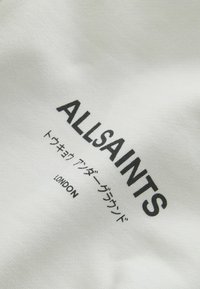 Tissu blanc avec texte noir indiquant "ALLSAINTS", avec des caractères japonais supplémentaires et "LONDON" en dessous. Texture lisse.