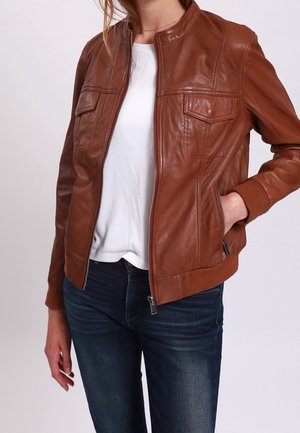Femme portant une veste en cuir marron par-dessus une chemise blanche et un jean bleu foncé, une main dans la poche de la veste et la fermeture éclair partiellement ouverte.
