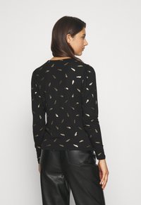 Blusa preta de manga longa com padrão de folhas douradas, apresentando um decote redondo e ombros ligeiramente franzidos, combinada com calças com aspeto de couro.