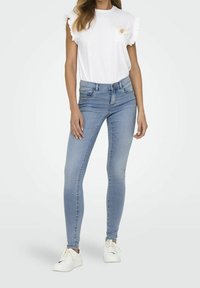 Světle modré skinny džíny s pěti kapsami a zipem, kombinované s bílým topem s krátkým rukávem, který má volánkové rukávy a kapsu na prsou.