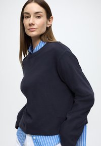 Námornícký sveter so štruktúrovaným prevedením, spustenými ramenami a okrúhlym výstrihom, nosený cez modrú a bielo pruhovanú košeľu.