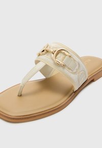 Sandales plates couleur crème avec une sangle texturée, des accents en métal doré et une semelle en caoutchouc marron, présentant un design de tongs pour un maintien sécurisé.