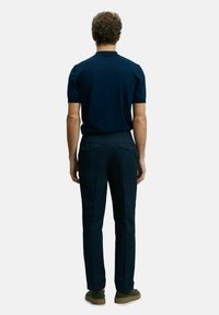 Polo shirt bleu marine à manches courtes, associé à un pantalon sombre avec poches arrière et jambes droites, complété par des chaussures vertes.