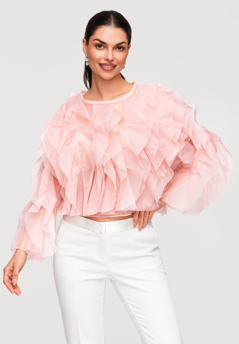 Jeune femme souriante, vêtue d'un haut rose transparent à volants superposés et d'un pantalon blanc, posant avec une main sur la hanche sur un fond uni.