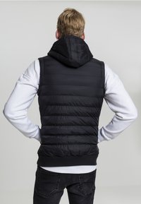 Vestă puffer neagră cu glugă, având o textură matlasată și un tiv cu nervuri, purtată peste un pulover alb, prezentată din spate.