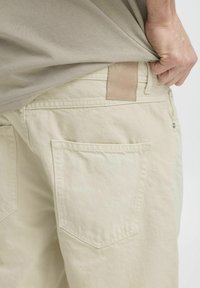 Beige Baumwollhosen verfügen über zwei Gesäßtaschen, ein Lederpatch mit Markenprägung und gestickte Details entlang des Taillenbunds.