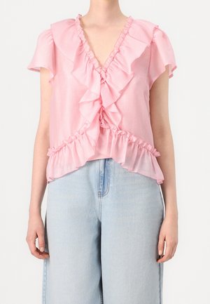 Blouse rose avec encolure et manches à volants, présentant un design cache-cœur. Tissu léger avec une texture douce. Associée à un jean en denim bleu clair.