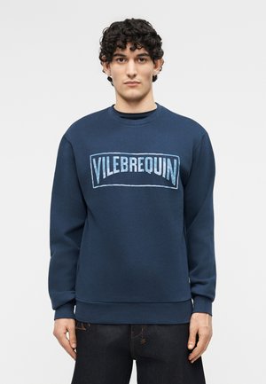 Άνδρας με σγουρά μαλλιά που φοράει μπλε σκούρο φούτερ με το λογότυπο "VILEBREQUIN", στέκεται απέναντι σε απλό ανοιχτό γκρι φόντο.