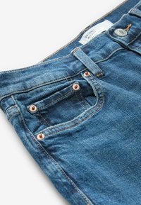 Dunne blauwe denim jeans met een middelhoge taille, koperen klinknagels, een klein voorvak en een label met de tekst "SKINNY MID RISE." Duurzame textuur.