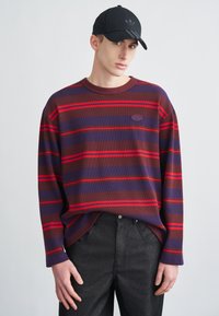 Ριγωτό πουλόβερ με μακριά μανίκια σε σκούρο μωβ, κόκκινο και καφέ με ριμπ υφή. Φορεμένο με μαύρο παντελόνι και μαύρο καπέλο με λογότυπα.