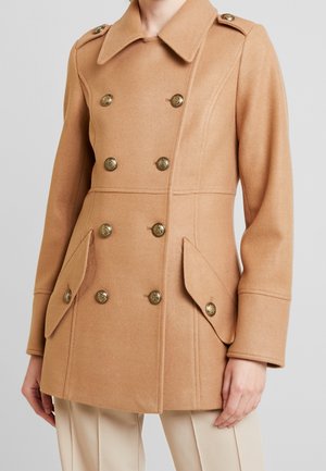 Kvinde iført en kamelfarvet dobbeltradet pea coat med guldknapper og klaplommer, kombineret med beige bukser.