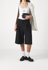 Czarne plisowane szorty, biały top na ramiączkach z logo, paskowany blezer, czarna torebka oraz czarne mokasyny na grubej podeszwie w połączeniu z białymi skarpetkami.