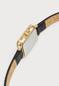 Montre rectangulaire avec un boîtier en acier inoxydable doré, un bracelet en cuir noir et un cadran de couleur argentée. Elle dispose d'une couronne et d'une gravure subtile.