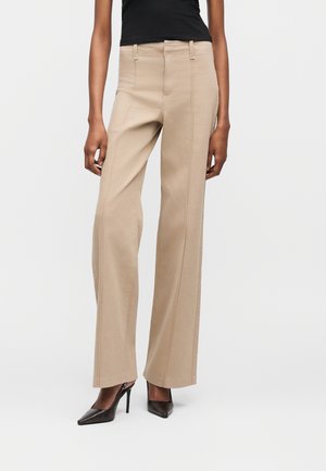 Person iført beige bukser med vide ben og sømme foran, sort ærmeløs top og sorte spidse højhælede sko mod hvid baggrund.