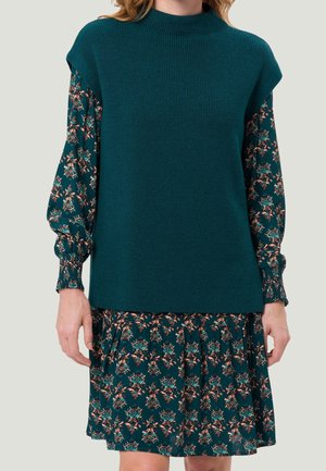 Kvinde iført en tealgrøn ærmeløs strikket vest over en langærmet blomsterkjole med teal, pink og beige blomsterprint.