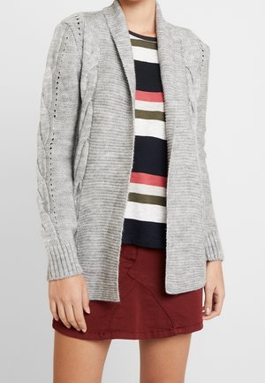 Kobieta w szarym otwartym kardiganie z rękawami z grubym splotem, w paskowaną koszulę w kolorach granatowym, białym, oliwkowym i koralowym oraz w rdzawą spódnicę.