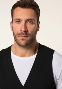 Chaleco negro sin mangas sobre una camisa blanca, con un diseño de escote en forma de V profundo y una textura de tela suave. Tonos de piel visibles en el cuello.