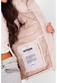 Beige puffersjakke med en shiny overflade, der har et trykt logo indeni, trykknapper og en etiket med plejeinstruktioner.