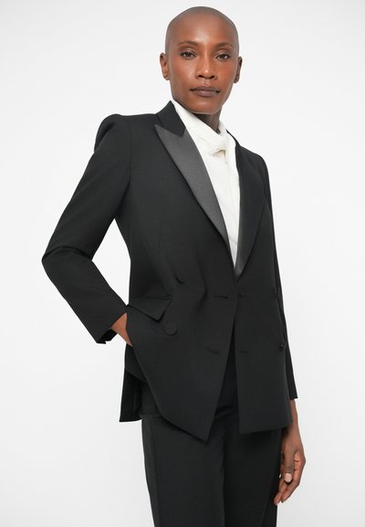 Blazer noir croisé avec des revers en satin, épaules structurées et deux boutons. Associé à une chemise blanche, il présente une coupe ajustée.