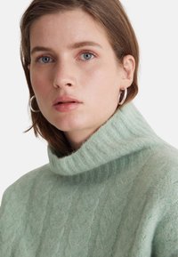 Grüner Pullover mit Zopfmuster und hohem, geripptem Kragen. Weiche Textur und lockere Passform, mit einem dezenten Muster und minimalen Details.