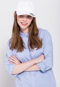 Hvid baseball cap båret med en lyseblå skjorte med knaplukning. Skjorten har oprullede ærmer, og modellen smiler med armene krysset.