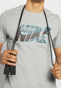 Gray t-shirt with a camouflage "NIKE" logo in teal and dark brown, holding a black jump rope with foam handles.