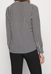 Sort og hvid houndstooth-mønstret bluse med lange ærmer, glat stof, afrundet kant, tætsiddende silhuet og knaplukning ved ærmerne.