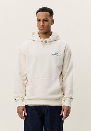 Junger Mann trägt einen cremefarbenen Hoodie mit dem Logo "Les Deux Society" auf der Brust und dunkelblaue Jeans und steht vor einem schlichten hellen Hintergrund.