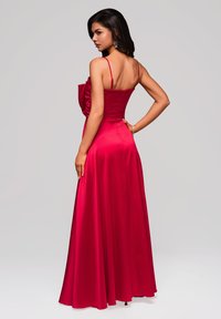 Lungo vestito di raso rosso con spalline sottili e corpetto aderente. Presenta una gonna fluida e un'accenno arricciato su un lato. Texture liscia in tutto il vestito.